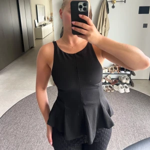 Svart insvängd topp med rosett i ryggen - Säljer en svart insvängd topp med breda axelband och snygg volang nertill. Toppen har öppen rygg med en rosett som knyts baktill. 