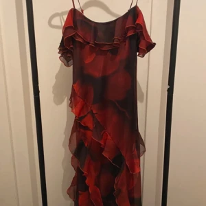 Röd volangklänning Rita Ora x Primark - Supersnygg röd och svart långklänning från Rita Ora x Primark med tunna axelband, volangdetaljer och offshoulder-look. Klänningen har ett flowigt tyg i polyester och ett subtilt mönster som ger en cool vibe. Perfekt för dig som vill sticka ut!