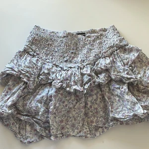 Blommig volangkjol från BikBok - Supersöt kort kjol från BikBok med blommigt mönster i lila, rosa och grå toner. Kjolen har bred smock i midjan och flera volanger som ger en flowy och lekfull känsla. Perfekt för vår och sommar när du vill ha en chill och feminin look.