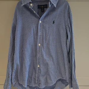 Blårutig skjorta från Ralph Lauren - Klassisk blårutig skjorta från Ralph Lauren med vit knappstängning och broderad logga på bröstet. Skjortan har lång ärm, krage och är tillverkad i mjuk bomull. Perfekt för dig som gillar en tidlös och stilren look.