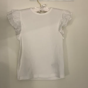 Vit volangtopp från Gina Tricot - Supersöt vit topp från Gina Tricot med volangärmar och rund halsringning. Toppen är ribbad och ärmlös med romantiska detaljer på axlarna. Perfekt för dig som gillar en feminin och trendig stil.