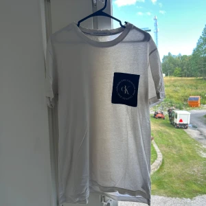 Vit t-shirt med blå ficka Calvin Klein - Vit t-shirt från Calvin Klein Jeans med en blå bröstficka och CK-logga. Klassisk rund halsringning och korta ärmar. Tunt och mjukt material som känns skönt mot huden. Perfekt för dig som gillar stilrena plagg med en twist.