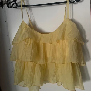 Gul volangig linnetopp med tunna axelband - Supersöt ljusgul linnetopp med flera lager volanger och tunna, justerbara axelband. Toppen är luftig och har en flowy känsla, perfekt för varma dagar. Materialet är lätt och ger en somrig vibe. Passar dig som gillar en lekfull och trendig stil.
