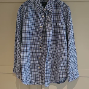 Rutig skjorta från Ralph Lauren blå/vit - Snygg rutig skjorta från Ralph Lauren i blått och vitt. Klassisk modell med knappar framtill och lång ärm. Skjortan har en avslappnad passform och är tillverkad i mjuk bomull. Perfekt för dig som gillar stilren och tidlös design.