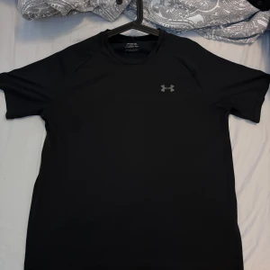 Svart tränings-tshirt från Under Armour - Svart t-shirt från Under Armour med diskret logga på bröstet. Tillverkad i ett lätt och snabbtorkande syntetmaterial, perfekt för träning. Klassisk passform med korta ärmar och rund hals. Enkel och stilren design som funkar till alla sportiga outfits.