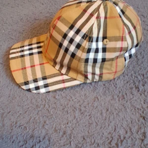 Burberry rutig keps i bomull - Snygg keps från Burberry i klassiskt beige, svart, vit och röd rutigt mönster. Justerbar rem bak med gulddetalj och ikonisk Burberry-logga på insidan. Perfekt accessoar för att lyfta din outfit med lyxig känsla.