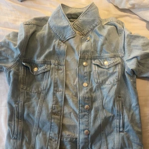 Ljusblå jeansjacka med volangdetalj - Säljer en ljusblå jeansjacka med snygga silvriga knappar och två bröstfickor framtill. Jackan har en unik volangdetalj på ryggen som ger en extra touch. Klassisk krage och raka sömmar, perfekt för dig som vill ha en chill men ändå trendig look.