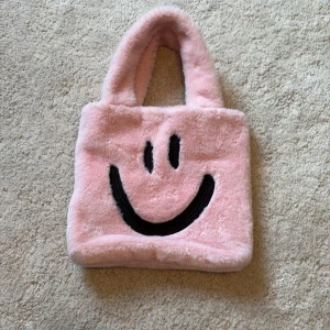 Rosa fluffig handväska från Lindex - Supergullig rosa handväska från Lindex i mjuk päls med en smiley på framsidan. Den har två handtag och även en axelrem som man ser på bilden. Den är lite sliten jämfört med själva väskan men kan även tas av🥰💓