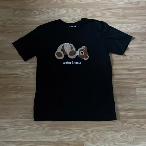 Svart Palm Angels t-shirt med björn - Svart t-shirt från Palm Angels i 100% bomull med ikoniskt broderat björnmotiv på bröstet och vit logga under. Klassisk passform och korta ärmar, perfekt för dig som vill sticka ut med streetwear-vibe.