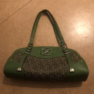 Grön DKNY axelväska med monogram - Snygg axelväska från DKNY i grönt skinn och textil med klassiskt DKNY-monogram. Väskan har silverfärgade metalldetaljer, dubbla handtag och dragkedja upptill. Unik form och coolt mönster gör den till ett statement-piece för din outfit.