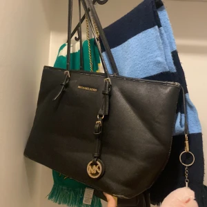 Svart axelväska Michael Kors skinn - Säljer en svart axelväska från Michael Kors i skinn med guldfärgade detaljer och MK-hänge. Väskan har dragkedja upptill, flera innerfack och långa axelband. Perfekt för dig som vill ha en stilren och rymlig väska med exklusiv känsla. Vara. Är ej äkta 