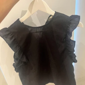 Svart blus med volangärmar - Säljer en svart blus med snygga volangdetaljer på axlarna. Toppen är ärmlös och har en lätt strukturerad yta som ger en cool känsla. Perfekt att styla med jeans eller kjol för en trendig look.