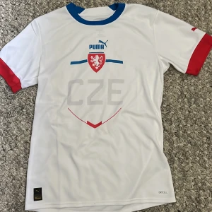 Tjeckien fotbollströja Puma S - Vit Tjeckien fotbollströja från Puma med blå krage och röda ärmslut. Framsidan har CZE-tryck, landslagets emblem och Puma-logga. Tillverkad i lätt och ventilerande polyester med DryCell-teknologi. Tjeckiska flaggan på ryggen.