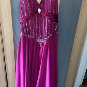 Lång ceriserosa balklänning med paljetter - Säljer en lång ceriserosa balklänning med glittriga paljetter och pärlor på livet. Klänningen har v-ringning, markerad midja med drapering och en elegant slits. Dragkedja bak för enkel påklädning. Perfekt för bal eller festligheter!