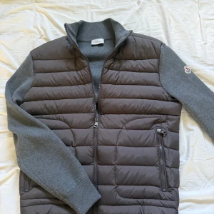 Grå pufferjacka från Moncler, M - Snygg grå pufferjacka från Moncler med stickade ärmar och quiltad front. Jackan har dragkedja framtill, två fickor med dragkedja och klassisk Moncler-logga på ärmen. Perfekt för dig som vill ha en stilren och sportig look.