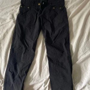 Svarta ridbyxor från Mountain Horse - Svarta ridbyxor från Mountain Horse med förstärkta partier på insidan av benen och guldiga knappar. Klassisk passform med bälteshällor och dragkedja. Perfekta för dig som vill ha snygga och funktionella byxor till stallet.