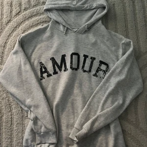 Grå hoodie med tryck 'AMOUR' och stjärna - Snygg ljusgrå hoodie med svart text 'AMOUR' framtill och en cool stjärna på ryggen. Tröjan har huva med snörning och ribbade muddar vid ärmslut och nederkant. Perfekt för en avslappnad och trendig look.