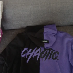 Svart och lila hoodie från Studio - Säljer en oversized hoodie från Studio i storlek L. Tröjan är delad i svart och lila med coolt tryck 'CHAOTIC' framtill. Den har huva och är tillverkad i mjuk bomull, perfekt för en avslappnad streetstyle-look.