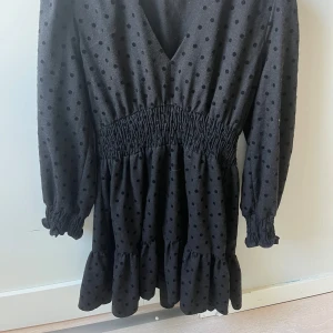 Svart prickig kortklänning från Zara - Supersöt svart kortklänning från Zara med prickigt mönster, v-ringning och smock i midjan. Klänningen har långa ärmar med resår vid ärmslut och volang nertill. Perfekt för dig som gillar en lekfull och trendig stil.