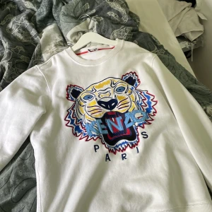 Vit Kenzo sweatshirt med tigerbrodyr - Kenzo sweatshirt i vitt med ikonisk broderad tiger i blått, gult, rött och svart framtill. Stora broderade bokstäver och Paris-tryck under motivet. Klassisk rund hals och långa ärmar. Perfekt statement-plagg för dig som gillar streetstyle.