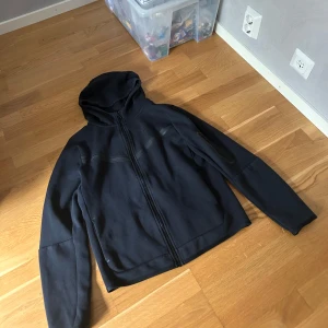 Svart Nike tech storlek 158-170  - Svart Nike tech med hel dragkedja framtill och diskret logga på bröstet. Jackan har huva, lång ärm och en praktisk ficka med dragkedja på ena ärmen. Tillverkad i mjuk bomullsmix, perfekt för en chill och sportig look.