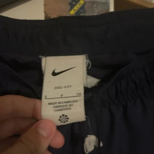 Nike PSG Dri-Fit träningsbyxor marinblå - Marinblå träningsbyxor från Nike med PSG-logga och orange swoosh. Byxorna är i Dri-Fit-material som andas och har resår i midjan med snörning. Perfekta för fotboll eller träning, med sportig look och bekväm passform.