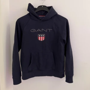 Mörkblå hoodie från GANT - Snygg mörkblå hoodie från GANT med broderad logga och sköld på bröstet. Klassisk känguruficka framtill och huva. Perfekt för dig som gillar stilren och sportig look. Mjuk bomullskänsla och långa ärmar. 