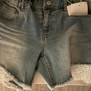 Jeansshorts med spetsdetalj - Snygga ljusblå jeansshorts med fransig kant och vit spets längs bensluten. Klassisk femficksmodell med knapp och dragkedja framtill. Perfekta för sommardagar när du vill ha en chill men ändå söt look.