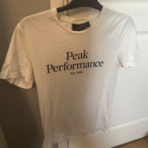 Vit Peak Performance t-shirt - Vit t-shirt från Peak Performance med svart tryck på bröstet där det står 'Peak Performance Est. 1986'. Klassisk rund halsringning. Tillverkad i mjuk bomull, perfekt för en clean och enkel stil. Nypris: 499kr
