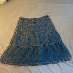 Vintage volangkjol med broderi  - Säljer en blå jeanskjol från KappAhl i storlek 38. Kjolen har volangdetaljer, två bakfickor med cool broderad söm och klassisk jeansknapp framtill. Perfekt för dig som gillar avslappnad och trendig stil.