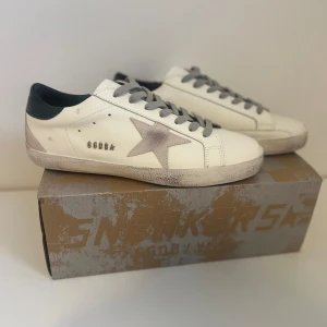 Golden Goose vita sneakers med stjärna - Snygga vita sneakers från Golden Goose med grå snörning och ikonisk grå stjärna på sidan. Skorna har svart hälparti och slitna detaljer på sulan för en cool vintage-look. Tillverkade i skinn och perfekta för dig som gillar streetstyle.