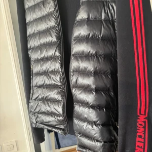 Svart dunväst från Moncler med röda detaljer - Snygg svart cardigan från Moncler med quiltad framsida och dragkedja. Köpt på Moncler i Milano. Ärmarna är svarta med tre röda ränder och Moncler-logga på ena ärmen. Materialet är polyester och dun, vilket gör den både lätt och varm. Kan gå ner lite i pris vid snabb affär 