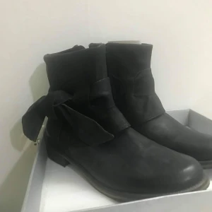 Svarta boots med rosett i skinn - Stilrena svarta boots från Ten Points i mjukt skinn med dekorativ rosett på sidan. Skorna har platt sula och rund tå, perfekt för dig som gillar en enkel men ändå unik look. Passar till många olika outfits och har en klassisk siluett.