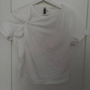 Vit t-shirt med twist från Divided - Vit t-shirt från Divided med kort ärm och cool twistad detalj vid axeln. Klassisk rund halsringning och rak passform. Perfekt för en clean och enkel look. Gjord i mjuk bomull som känns skön mot huden.