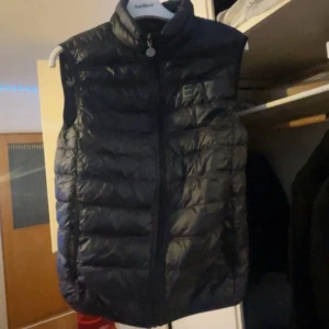 Svart dunväst från EA7 Emporio Armani - Säljer en svart dunväst från EA7 Emporio Armani med hög krage och diskret logga på bröstet. Västen har quiltad design och dragkedja framtill. Perfekt att slänga över en hoodie för en chill look.