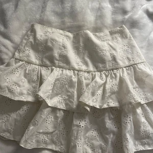 Vit volangkjol med broderade blommor - Supersöt vit kortkjol med broderade blommor och volanger. Kjolen har en romantisk känsla med sina lager och spetsdetaljer. Perfekt för sommarens alla tillfällen och lätt att matcha med en enkel topp.