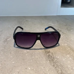 Carrera solglasögon - Snygga svarta solglasögon från Carrera med klassisk pilotform och mörka tonade glas. Nypris 799. Ramen är i svart plast med Carrera-logga på sidan. Perfekta för att ge en cool vibe till din outfit.