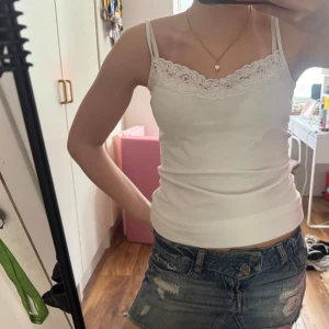 Vit spetslinnetopp med smala axelband - Supersöt vitt linne med smala axelband och snygg spetsdetalj längs urringningen. Toppen är figurnära och perfekt för varma dagar. Enkel att matcha med jeans eller kjol.