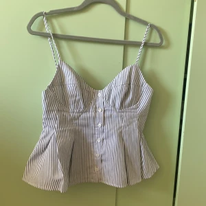 linnetopp med knappar - Supersöt linnetopp med smala axelband och knappar framtill. Toppen är vit med tunna blå ränder och har en figurnära passform upptill som blir lite utsvängd nertill. Perfekt för varma dagar och ger en fräsch, somrig vibe.