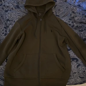 Mörkgrön hoodie från Polo Ralph Lauren - Snygg mörkgrön hoodie från Polo Ralph Lauren med dragkedja och huva. Klassisk broderad logga på bröstet, känguruficka framtill och justerbara snören i huvan. Perfekt för chill dagar och enkel att matcha med jeans eller joggers.
