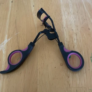 Svart och rosa ögonfransböjare - En ögonfransböjare i svart metall med detaljer i rosa gummi på handtagen och kudden. Ergonomiskt grepp med rundade handtag för enkel användning. Perfekt för att ge fransarna en snygg böj och öppna upp blicken.