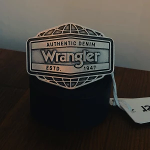 Svart Wrangler bälte med stor buckle - Skriv om du vill förhandla om pengarna 😁.Säljer ett par svarta bälte från Wrangler med en stor, silverfärgad buckle där det står 'Authentic Denim Wrangler ESTD. 1947'. Bältet är gjort i skinn och har en cool western-vibe. 