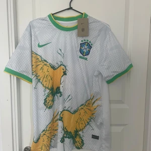 Vit Brasilien fotbollströja Nike L - Snygg vit Brasilien fotbollströja från Nike med gröna och gula detaljer. Tröjan har korta ärmar, grönt och gult ribbade muddar och krage, samt stora gula fåglar tryckta på framsidan. Tillverkad i Dri-FIT material som andas, perfekt för match eller träning.