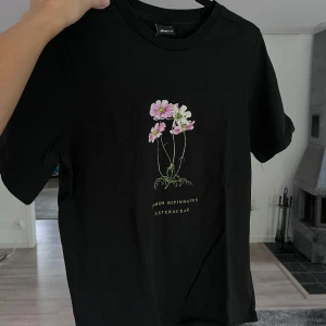 Svart t-shirt med blommor - Svart t-shirt från Gina Tricot med tryck av rosa och vita blommor på bröstet. Klassisk rund halsringning och korta ärmar. Tillverkad i mjuk bomull, perfekt för en chill och stilren look. Önskar du veta exakta mått? Tveka inte att höra av dig!
