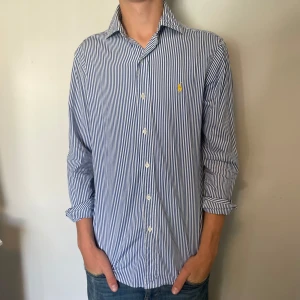Ralph lauren skjorta - Säljer denna riktigt snygga randiga Ralph lauren skjorta i vitt och blått med guldfärgat märke, den är i mycket bra skick utan några defekter, storlek S modell custom fit, den är 75 cm lång och 46 cm bred under armhålorna, hör gärna av dig vid frågor!