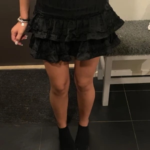 Svart volangkjol med broderi - Supersöt svart kortkjol med flera lager volanger och broderade detaljer. Kjolen har en diskret dragkedja i sidan. Perfekt  för dig som vill ha en trendig och lekfull look.