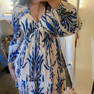 Vit och blå mönstrad kortklänning - Superfin kortklänning med blått blommönster på vit botten. Klänningen har v-ringning, långa ärmar och ett luftigt, plisserat tyg som ger en flowy känsla. Perfekt för dig som gillar att sticka ut med färg och mönster. Helt slutsåld 