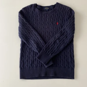 Mörkblå kabelstickad tröja Polo Ralph Lauren - Kabelstickad mörkblå tröja från Polo Ralph Lauren med klassisk röd broderad logga på bröstet. Tröjan har rund halsringning, långa ärmar och breda ribbade muddar. 