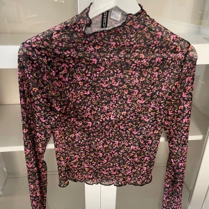 Blommig mesh-top från H&M Divided - Säljer en transparent långärmad mesh-top från H&M Divided med svart botten och små rosa blommor över hela. Toppen har en tight passform och vågig kant nertill, perfekt att styla lager på lager eller ha som en elegant blus/topp 
