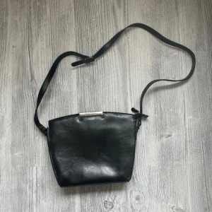 Svart axelväska från Puccini i skinn - Stilren svart axelväska från Puccini i äkta skinn med justerbar rem och silverfärgad metall-detalj upptill. Väskan har flera fack inuti, inklusive dragkedjefack, perfekt för att hålla ordning på dina grejer. Axelremmen är avklippt och blir max 105cm.